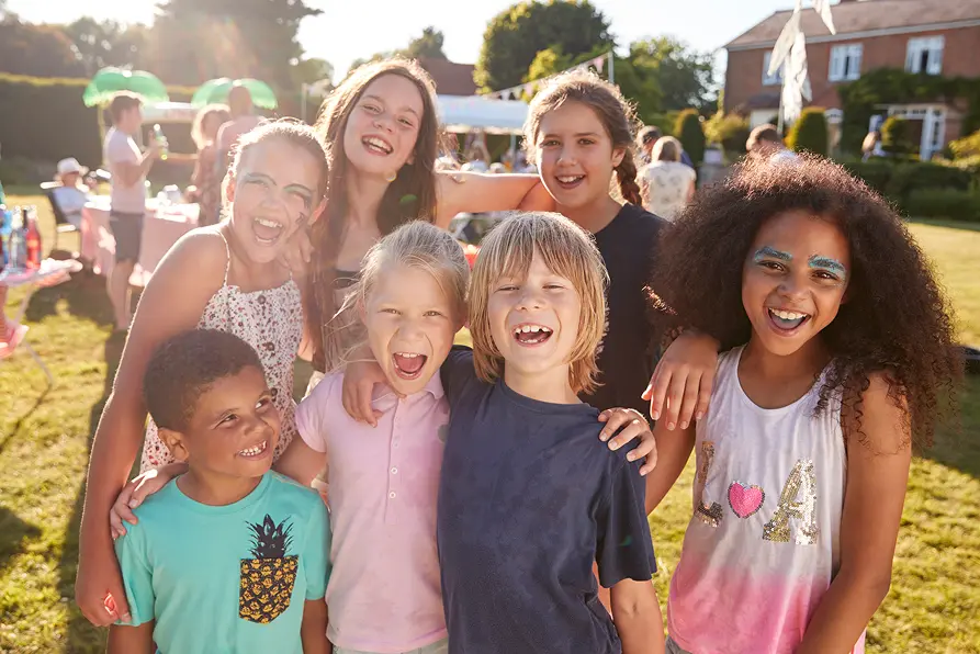 A group of six smiling children stand together outdoors on a sunny day.
