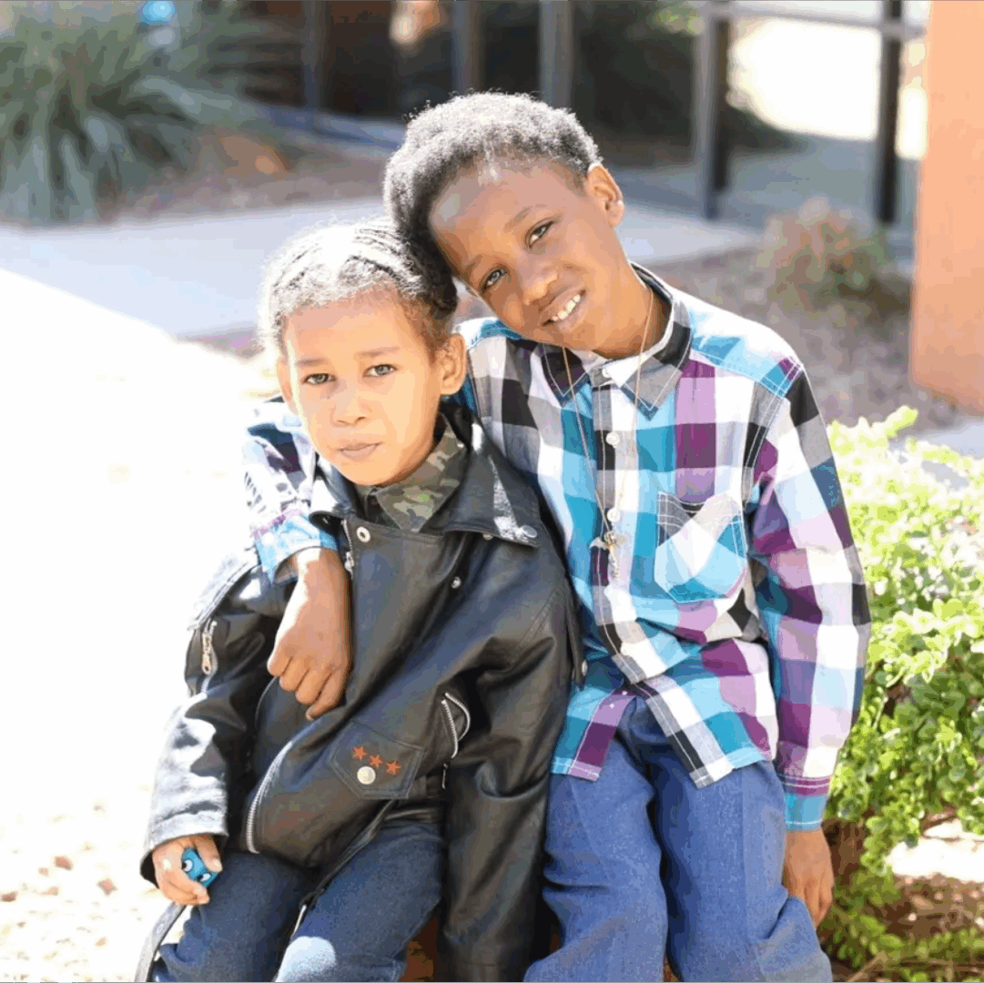 Two children sitting together, smiling outdoors.