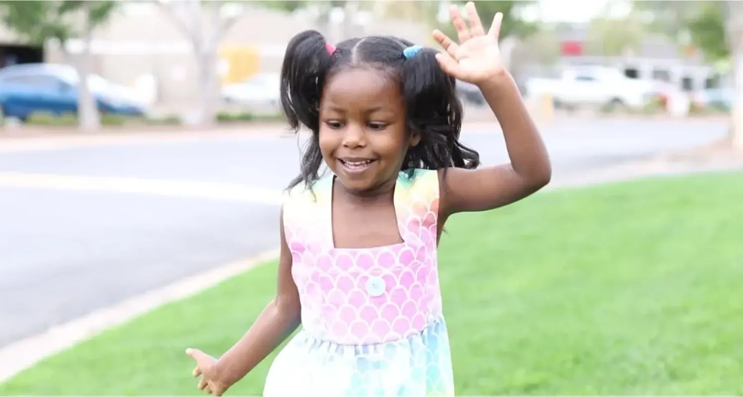 Girl smiling and waving on grassy area.